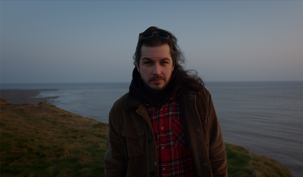 Image of a man with a brown jacket standing on a cliff top by the sea. 