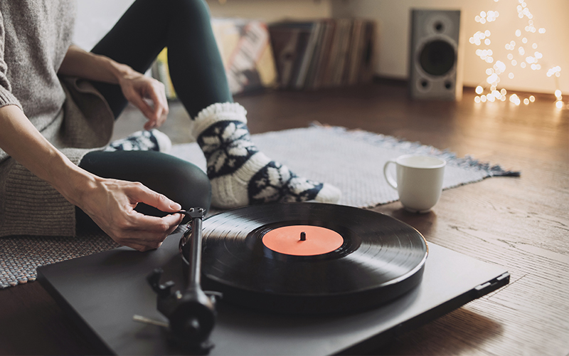 A woman sat on a rug on a wooden floor placing the tone arm onto the vinyl record. 