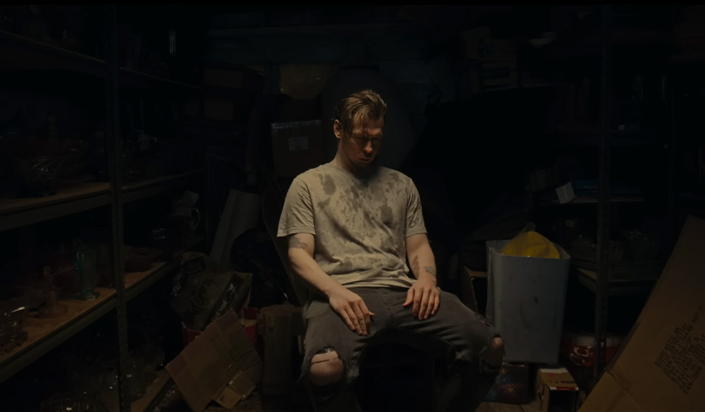 Man with a stained beige t-shirt sitting on a chair in a moody garage setting. 