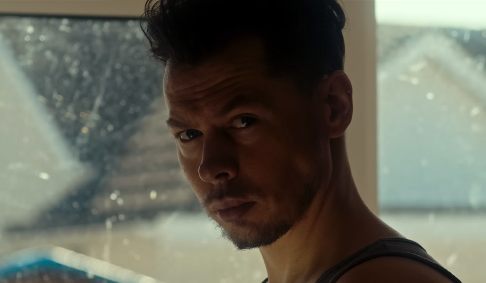 Still image of a man looking directly at the camera stood in front of a window. 