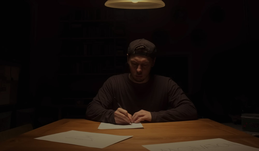 Picture of a man with a backwards cap sitting at a desk with a pen and paper, with a soft overhead light fixture above. 