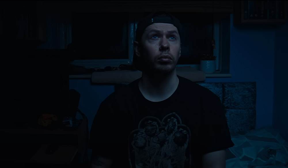 A still image of a man with a black t-shirt sitting in a dark bedroom. 