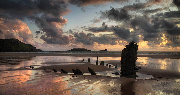 Landscape photo of the Welsh coast at sunset with a wrecked vessel in the foreground.