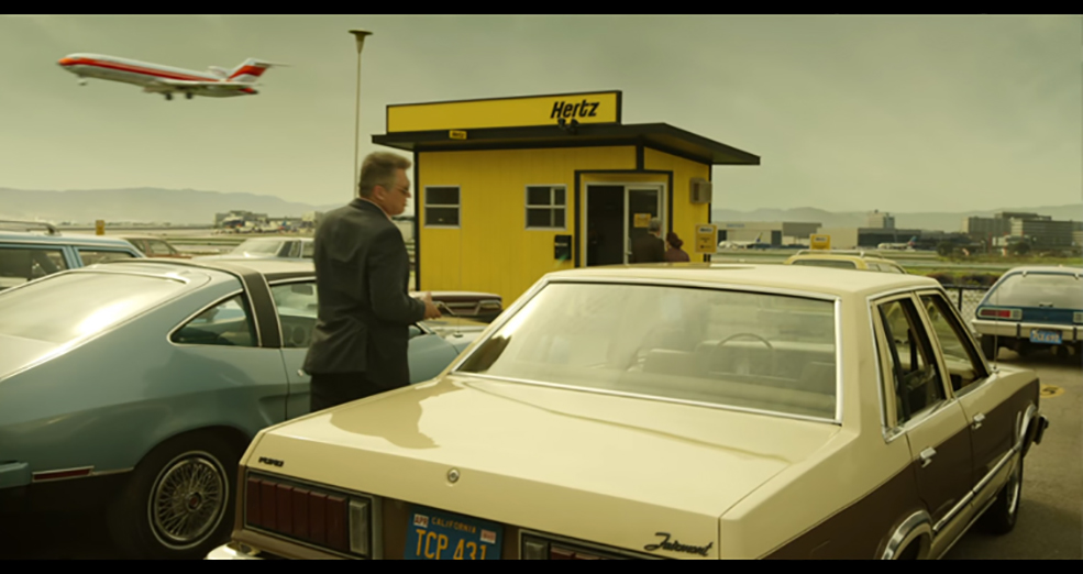 Still image of a man walking in between two cars from the film Mindhunter with an airplane in the sky 