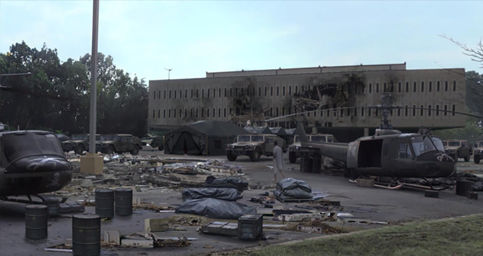 Image of a scene consisting of wrecked helicopters, military trucks and tents, a large damaged building with a lone male wanderer amongst the scene. 