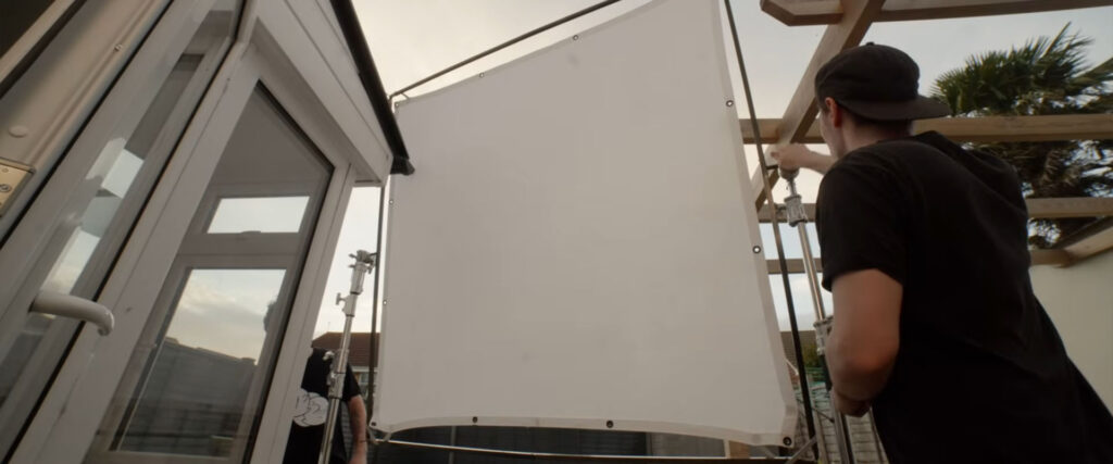 A person in a cap is adjusting a large white diffuser panel outside, next to an open glass door with tropical plants in the background