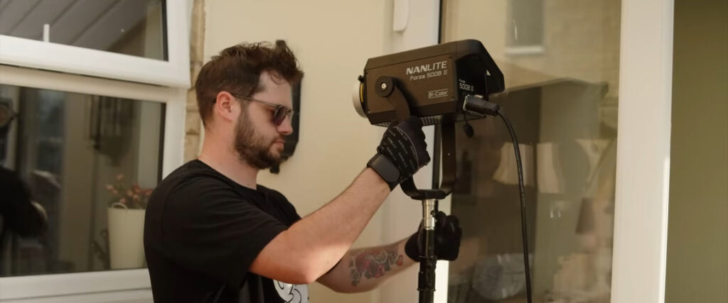 A bearded man in sunglasses adjusts a "NANLITE Forza 500B II" lighting equipment near a window.