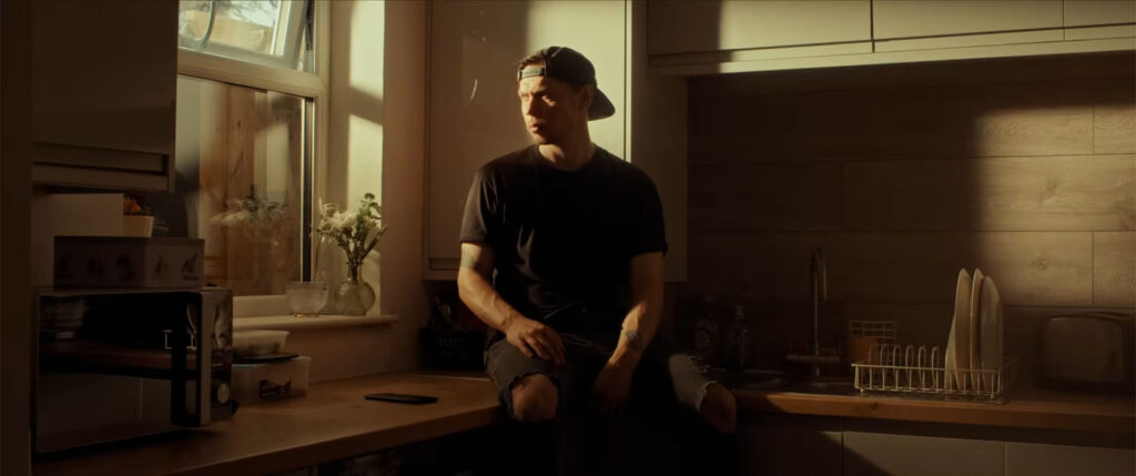 A man in a black t-shirt and ripped jeans sits on a counter by a window, bathed in warm light. 